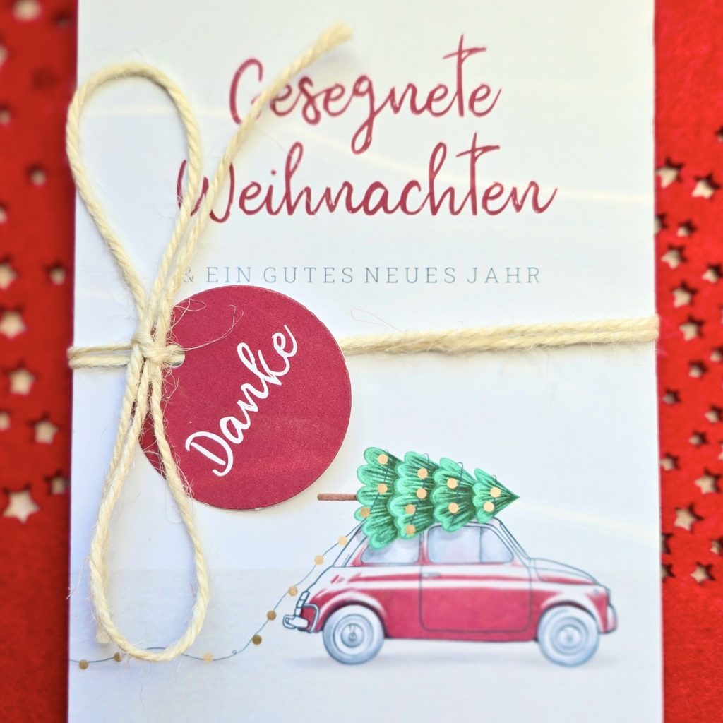 Das Bild zeigt eine Weihnachtskarte, die auf einem roten Untergrund mit Sternenmuster steht. Text auf der Karte: "Gesegnete Weihnachten und ein gutes neues Jahr." Die Karte ist mit einem Paketband zusammengebunden. Darauf klebt ein roter, kreisrunder Aufkleber, mit dem Wort "Danke". Unten auf der Karte ist ein Auto sichtbar. Auf dem Dach des Autos liegt ein geschmückter Weihnachtsbaum.