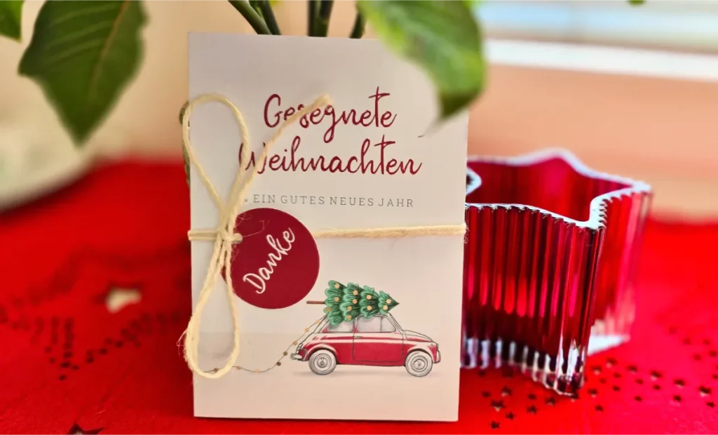 Das Bild zeigt eine Weihnachtskarte, die auf einem roten Untergrund mit Sternenmuster steht. Text auf der Karte: "Gesegnete Weihnachten und ein gutes neues Jahr." Die Karte ist mit einem Paketband zusammengebunden. Darauf klebt ein roter, kreisrunder Aufkleber, mit dem Wort "Danke". Unten auf der Karte ist ein Auto sichtbar. Auf dem Dach des Autos liegt ein geschmückter Weihnachtsbaum.